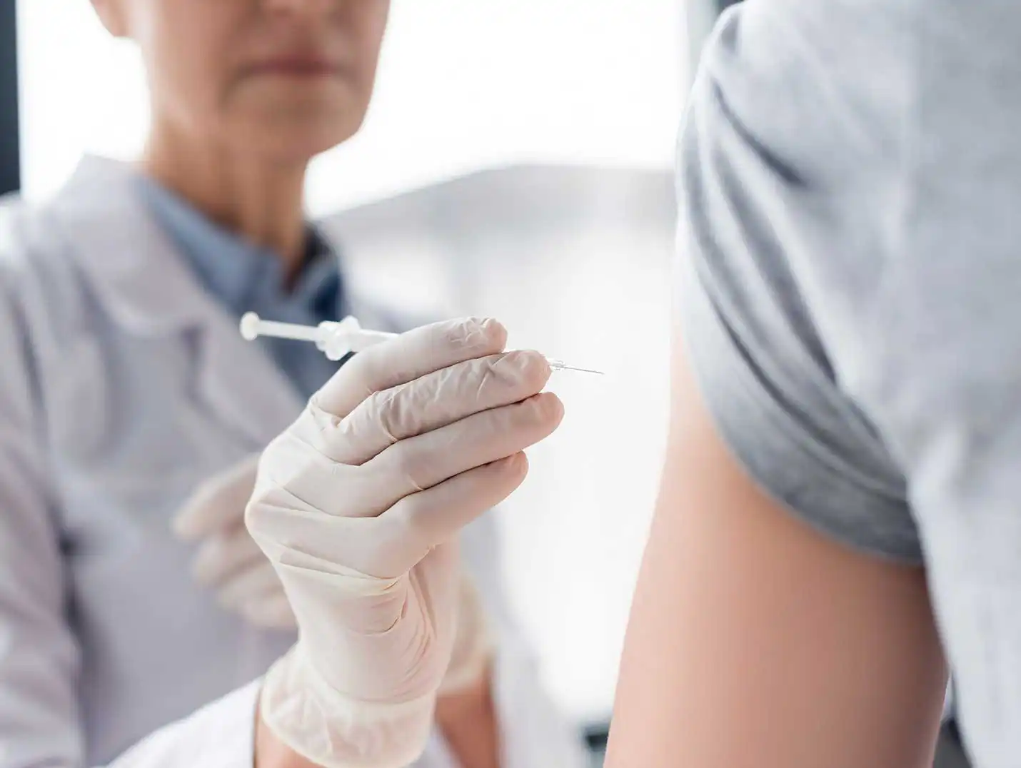 Médico con guantes blancos sosteniendo una jeringa para aplicar una inyección en el brazo izquierdo del paciente. 